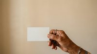 Male hand with medium-dark skin tone holding blank white business card against neutral beige background for branding and corporate identity mockup purposes.