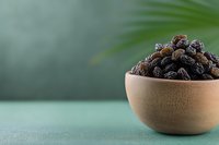 Fresh dark raisins in wooden bowl on green background with palm leaf, healthy dried fruit snack for natural sweet flavor and nutrition.