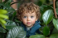Adorable curly-haired toddler boy with blue eyes peeking through lush green tropical plants in natural indoor setting for family lifestyle content.