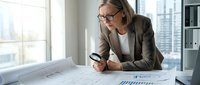 Professional mature Caucasian businesswoman architect examining blueprints with magnifying glass in modern office workspace for construction planning.