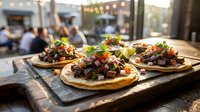 Gourmet tacos with seasoned meat, fresh toppings and cilantro served on wooden board at outdoor restaurant patio with blurred guests and string lights in background.