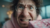 Excited middle-aged Caucasian man with glasses and messy hair holding gift box with wide-eyed surprised expression in blurred indoor setting.