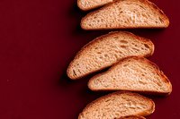 Fresh sliced sourdough bread arranged in diagonal pattern on dark red background, showcasing texture and golden crust. Food photography for bakery marketing.