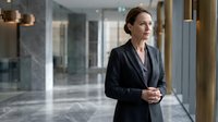 Professional middle-aged Caucasian businesswoman in dark suit standing confidently in modern corporate office lobby with marble walls and glass elements.