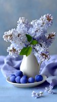 Delicate lilac flowers in white ceramic vase with fresh blueberries on plate, arranged on soft blue fabric for spring home decor.
