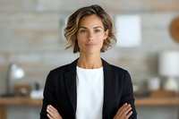 Young professional woman in black blazer and white top with confident pose, arms crossed, standing in modern office interior with soft natural lighting.