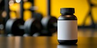 Black supplement bottle with blank white label on blurred gym background with dumbbells and yellow wall, mockup for fitness nutrition products.