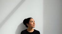Young woman with dark hair in a bun, South Asian ethnicity, wearing black shirt, looking upward thoughtfully against white wall with natural shadow lighting for contemplation or aspiration concepts.