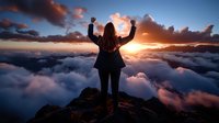 Young woman with raised arms standing on mountain peak above clouds at dramatic sunset, expressing freedom and achievement in nature. Success concept.
