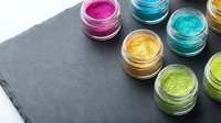 Colorful glitter and shimmer powders in open containers arranged on dark slate surface, isolated on white background for cosmetics, makeup, and design projects.