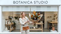 Woman florist holding autumn flower bouquet in modern botanical studio storefront with natural wood shelving and potted plants displayed.