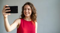 Happy young Caucasian woman in red sleeveless top taking selfie with smartphone, smiling at camera against neutral background for social media content.