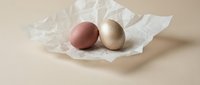 Two elegant Easter eggs in rose gold and champagne metallic finish resting on crumpled white tissue paper against neutral beige background for holiday decor.