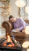 Young woman in purple sweater writing in notebook on leather armchair by fireplace with warm fairy lights creating cozy winter atmosphere at home.