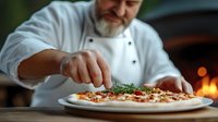 Caucasian male chef in white uniform garnishing fresh pizza with herbs, wood-fired oven with flames in background. Professional culinary preparation.