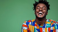 Young African American man with dreadlocks laughing joyfully wearing colorful geometric patterned shirt against green studio background, expressing genuine happiness.