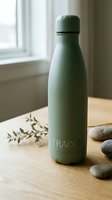 Sage green insulated water bottle with metal band on wooden table near window, surrounded by stones and dried botanicals for eco-friendly lifestyle and wellness product photography.