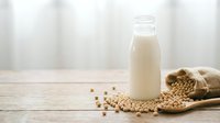 Fresh soy milk in glass bottle with raw soybeans scattered on wooden table, healthy plant-based dairy alternative for vegan nutrition and cooking.