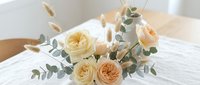 Elegant cream and peach rose bouquet with eucalyptus and dried grasses on white bedding, soft natural lighting for wedding decor and romantic occasions.