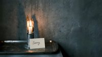 Lit candle on a metal tray beside a handwritten sorry note against a textured gray wall, evoking remorse, apology, and quiet emotion.
