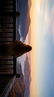 Brown horse standing by wooden fence at golden hour sunset, gazing toward distant mountains and glowing sky. Suitable for equestrian, nature, and travel content.