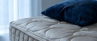Comfortable mattress with quilted surface topped with soft navy blue pillow in bright bedroom setting with natural light from window.
