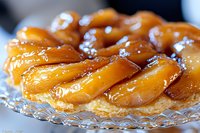 Caramelized pear tart on crystal plate features golden-brown fruit arranged in concentric circles atop flaky pastry base, glazed with sweet syrup.