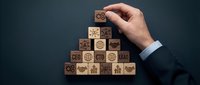 Young adult male hand in business suit placing wooden cube with organizational chart icon atop pyramid structure of blocks displaying corporate symbols, representing hierarchy, leadership, and strategic planning.