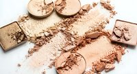 Broken makeup powder compacts and eyeshadows scattered with crumbled cosmetic dust on white background for beauty and fashion design projects.