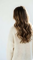 Young woman with long wavy brown hair wearing cream knit sweater from behind against neutral background for hair care and fashion concepts.