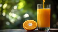 Fresh orange juice being poured into glass with orange half on wooden table against blurred green nature background for healthy breakfast concept.