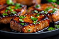 Grilled pork chops glazed with honey soy marinade on black plate, garnished with fresh chopped parsley. Close up view showing juicy meat texture and caramelization.