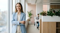 Professional businesswoman in light blue blazer standing confidently in modern office with arms crossed, smiling at camera in corporate workplace environment.