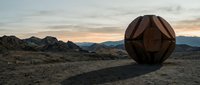 Geometric metal sphere sculpture in desert landscape at sunset with mountains and rocky terrain in background.