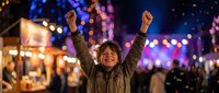 Excited young boy raising fists in triumph at a vibrant night festival, colorful bokeh lights and festive crowd in the background.