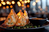 Crispy golden fried wontons with dipping sauce on dark plate garnished with chopped green onions. Blurred bokeh lights create warm restaurant ambiance.