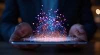 Magical glowing particles rising from digital tablet screen held by person in dark room, creating ethereal light display of blue and purple sparkles.