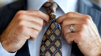 Close-up of male hands adjusting elegant blue and gold patterned silk necktie, wearing wedding ring and formal business attire, showing professional grooming.