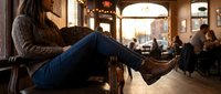 Young woman in cozy sweater and jeans relaxing in warm cafe with soft lighting and blurred customers in background creating intimate atmosphere.