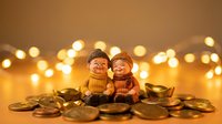 Cute ceramic figurines of elderly couple sitting together surrounded by golden coins with warm bokeh lights creating cozy atmosphere for wealth concept.