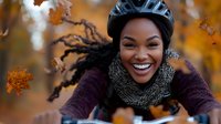 Young African American woman cyclist smiling while riding bike in autumn park, wearing protective helmet and warm knitted scarf against orange fall foliage background.
