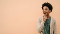 Young man with dark curly hair and medium brown skin tone wearing casual cardigan and t-shirt, smiling with hand near chin against neutral beige background for portrait and lifestyle use.