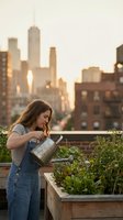 Young woman watering plants in urban rooftop garden at sunset with city skyline background for sustainable living and gardening concepts.