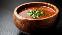 Spicy red soup with fresh green herbs garnish served in rustic wooden bowl on dark slate surface for culinary and food styling projects.