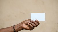 Close-up of a person with dark skin tone holding a blank white card or paper against a neutral beige background, suitable for mockups and promotional designs.