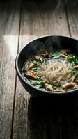 Steaming bowl of mushroom noodle soup with fresh herbs in dark ceramic bowl on rustic wooden table background for comfort food and healthy dining concepts.