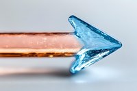 Blue crystal arrow shape on copper metal rod against light background, macro photography showing transparent mineral structure and light refraction.