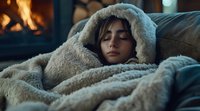 Young Caucasian woman wrapped in cozy blanket relaxing by fireplace on winter evening, eyes closed in peaceful rest. Warm atmospheric lighting creates tranquil mood.
