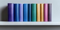 Colorful book spines arranged in rainbow order standing on white shelf against gray wall, creating minimalist gradient pattern from purple through blue to pink.