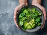 Hands holding bowl with fresh green salad, sliced avocado, spinach leaves and microgreens on gray background, healthy vegan food concept.